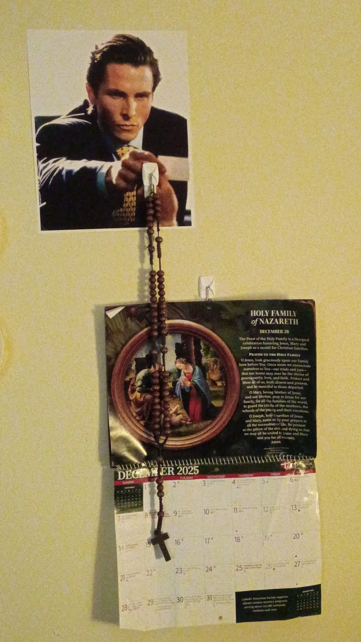 my poster of patrick bateman. there is a rosary hanging from the wall positioned to seem like he is handing the rosary to you. there is a blank calendar set to december
